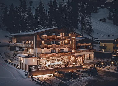 HOTEL TIROLER BUAM, Winteransicht Abend Beleuchtetes Hotel in winterlicher Berglandschaft in Saalbach mit Holzfassade, Terrasse und schneebedecktem Parkplatz