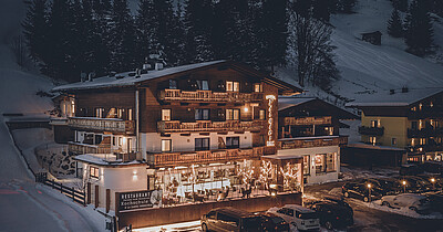 Beleuchtetes Hotel in winterlicher Berglandschaft in Saalbach mit Holzfassade, Terrasse und schneebedecktem Parkplatz