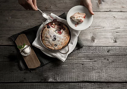 Anrichten von frischem Kaiserschmarren aus der Pfanne auf einem Holztisch mit Beeren, Puderzucker und Vanilleeis