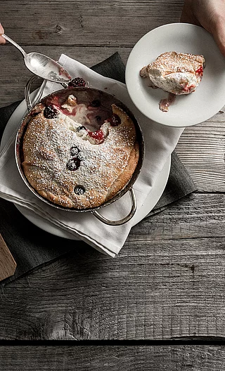 Anrichten von frischem Kaiserschmarren aus der Pfanne auf einem Holztisch mit Beeren, Puderzucker und Vanilleeis