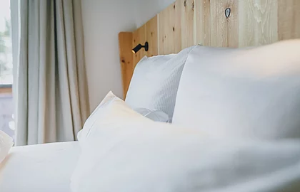Detailaufnahme eines Bettes mit hellem Holz-Kopfteil, weißen Kissen und beigefarbenem Vorhang
