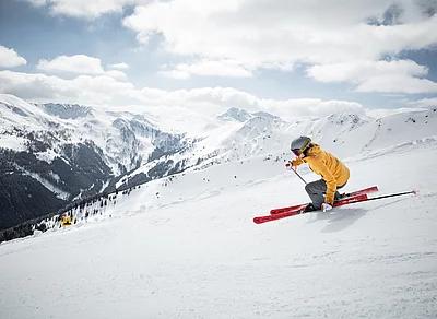 Skifahrerin in gelber Jacke fährt dynamisch eine tiefwinterliche verschneite Piste vor alpiner Bergkulisse hinab