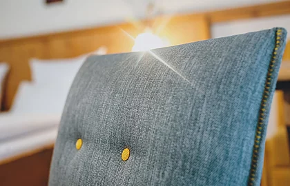 Detailaufnahme einer graublauen Stuhllehne mit gelben Knöpfen, im Hintergrund ein Bett mit Holzdesign