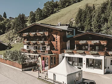 Sommerliche Außenaufnahme des Hotel Tiroler Buam Saalbach mit Blumenkästen an den Balkonen und sommerlichen Bergwiesen und Wald im Hintergrund