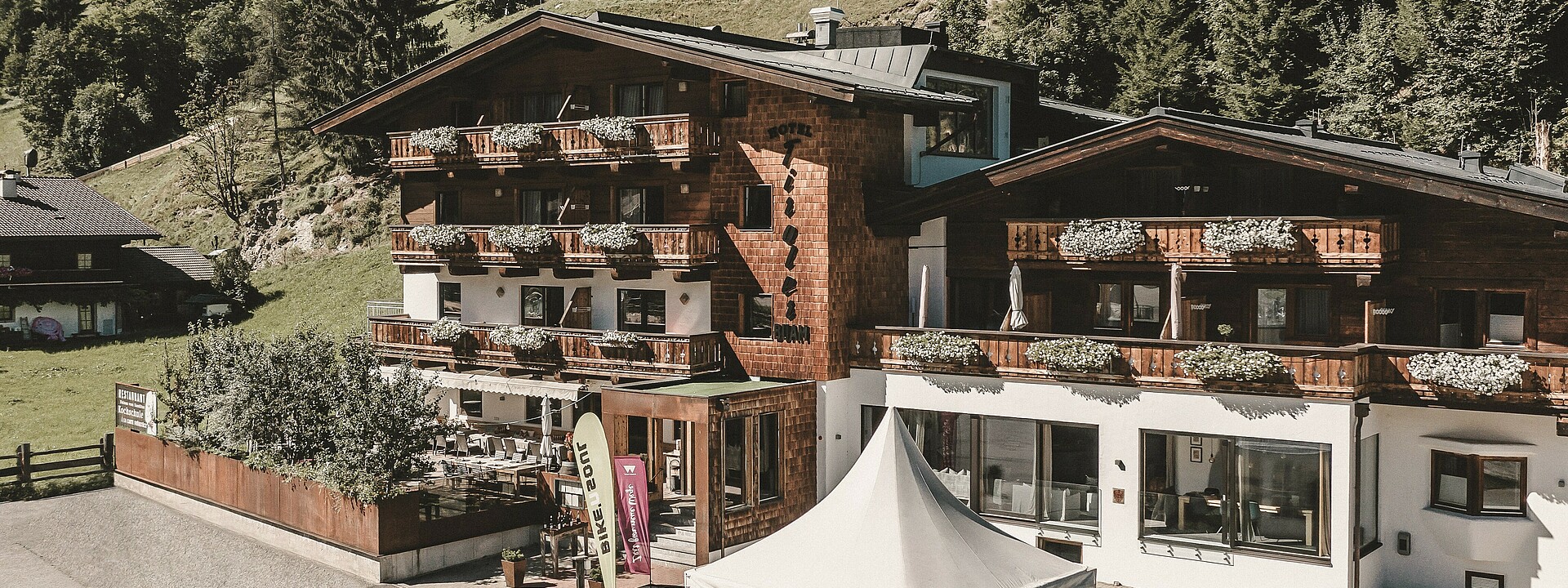 Sommerliche Außenaufnahme des Hotel Tiroler Buam Saalbach mit Blumenkästen an den Balkonen und sommerlichen Bergwiesen und Wald im Hintergrund