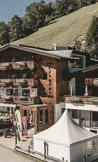 Sommerliche Außenaufnahme des Hotel Tiroler Buam Saalbach mit Blumenkästen an den Balkonen und sommerlichen Bergwiesen und Wald im Hintergrund