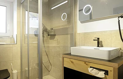 Modernes Badezimmer mit Glasdusche, beleuchtetem Spiegel, Waschtisch aus Holz und beigem Fliesendesign