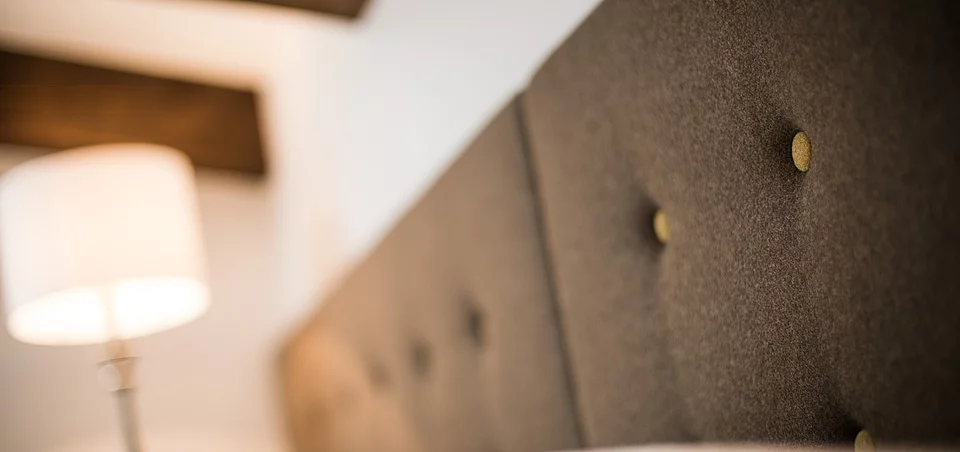 Zimmer & Appartments Detailaufnahme eines gepolsterten Betthaupts mit Knöpfen, daneben eine stilvolle Nachttischlampe