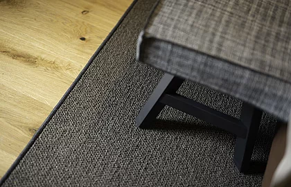 Detailaufnahme eines Stuhlbeins auf dunkelgrauem Teppich neben hellem Holzboden in einem Hotelzimmer
