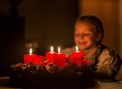 Adventkranz Kind Kind betrachtet lächelnd einen Adventskranz mit vier brennenden roten Kerzen in weihnachtlicher Atmosphäre
