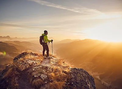 Wanderer in leuchtend gelber Jacke steht auf Bergspitze des Tristkogels, Sonnenaufgang beleuchtet die umliegenden Täler