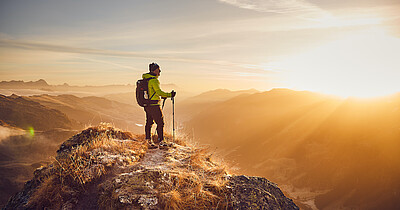 Wanderer in leuchtend gelber Jacke steht auf Bergspitze des Tristkogels, Sonnenaufgang beleuchtet die umliegenden Täler