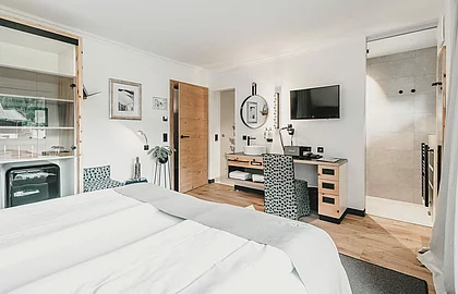 Helles Hotelzimmer mit Doppelbett, Holzmöbeln, Schreibtisch, Smeg-Kühlschrank und Einblick in ein modernes Badezimmer