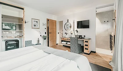 Helles Hotelzimmer mit Doppelbett, Holzmöbeln, Schreibtisch, Smeg-Kühlschrank und Einblick in ein modernes Badezimmer