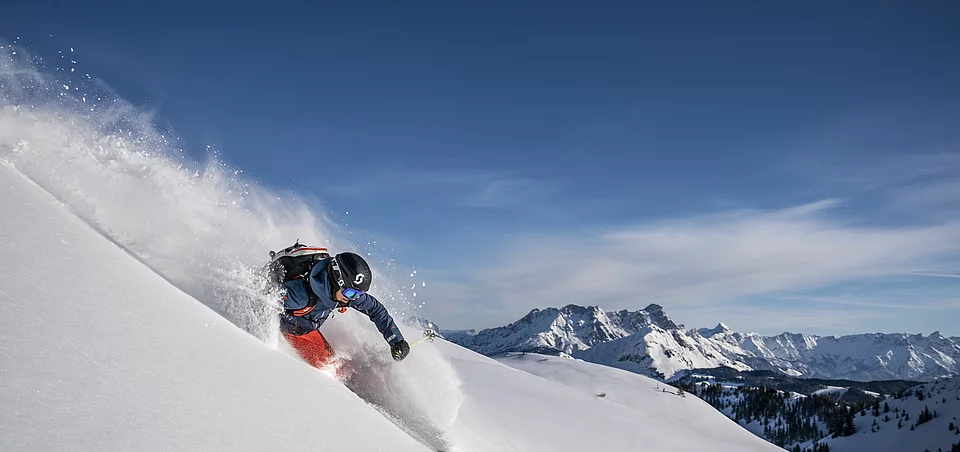 Ski In Ski Out ❄ Ski hotel Saalbach Freerider mit Helm und Skiern fährt dynamisch durch frischen Pulverschnee in alpiner Berglandschaft